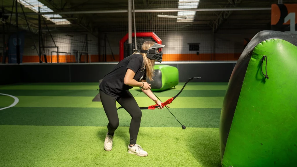 Personne jouant à l'activité Archery Tag, avec un arc dans la main, sur un terrain d'herbe synthétique