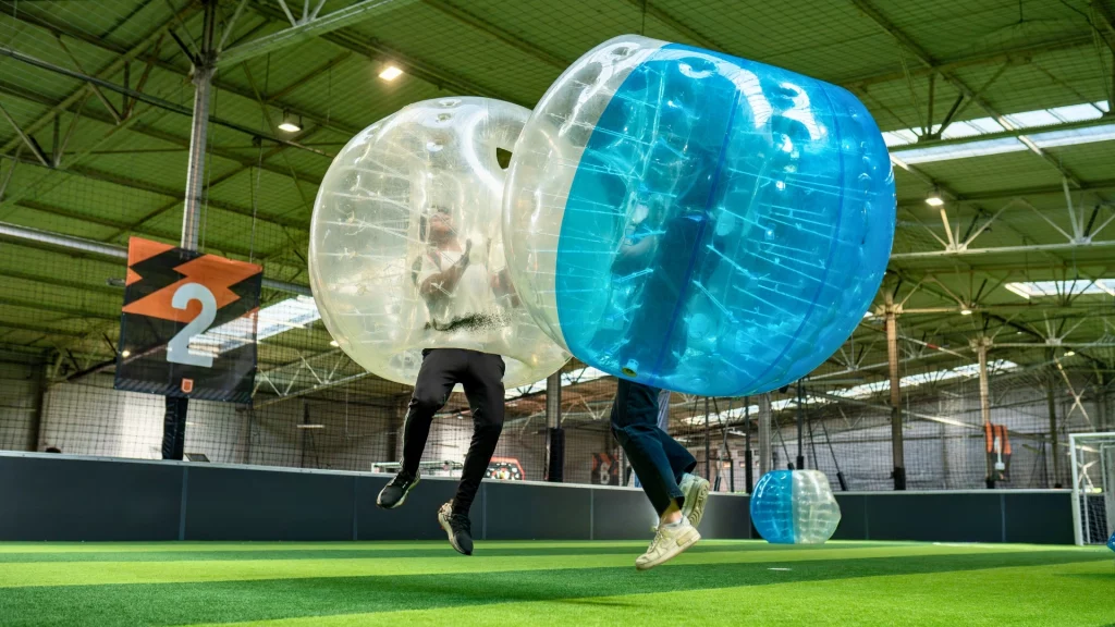 Deux personnes en bulles transparentes bleues et grises jouant au bubble football dans une salle couverte sur un terrain en gazon synthétique.